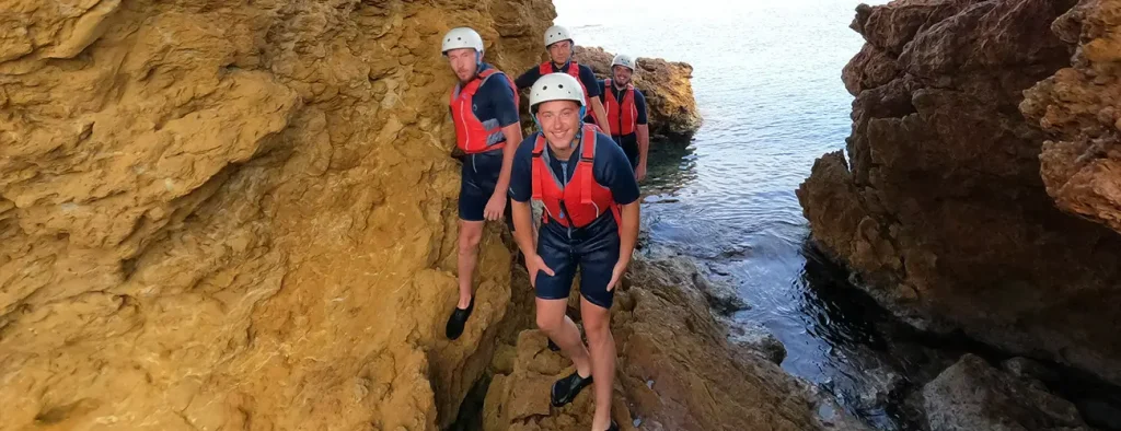 Grupo de cuatro hombres con cascos y chalecos salvavidas atravesando formaciones rocosas doradas durante una experiencia de coasteering en Ibiza.