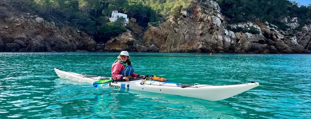 Mujer remando en un kayak de mar blanco a traves de aguas cristalinas turquesas con acantilados rocosos y vegetacion verde de fondo en Ibiza.
