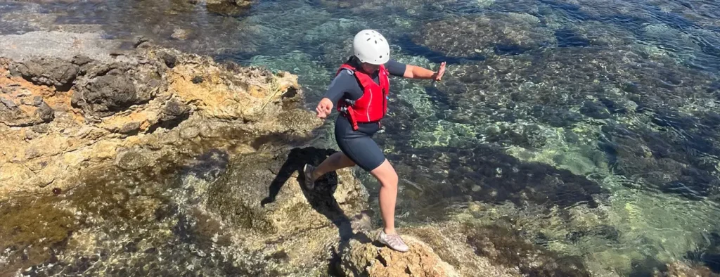 Participant wearing helmet and life jacket jumping from a golden rock into crystal clear waters during a coasteering session in Ibiza