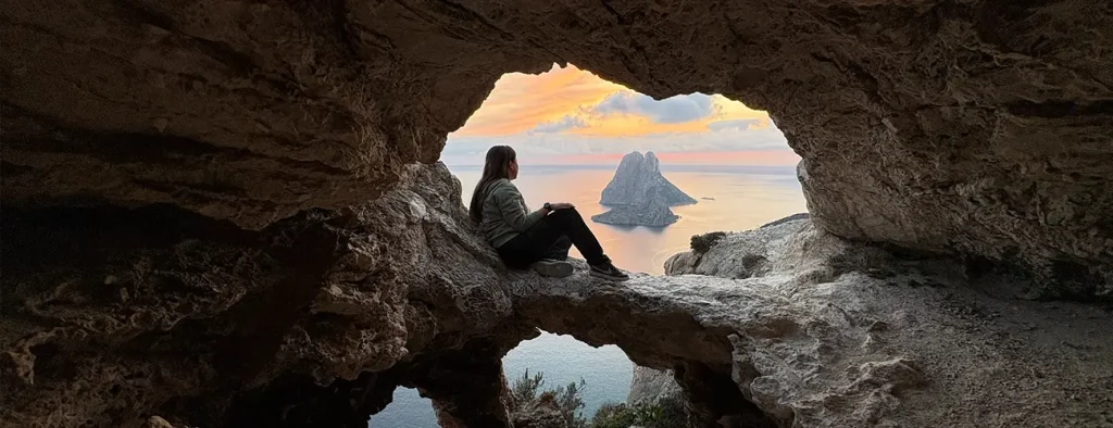 Woman sitting inside a natural rock cave watching the sunset over Es Vedra island with pink and orange sky colors in Ibiza
